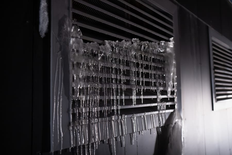 Air conditioner outdoor unit covered with thick ice buildup and icicles on metal vent grille, frozen cooling system detail in winter conditions