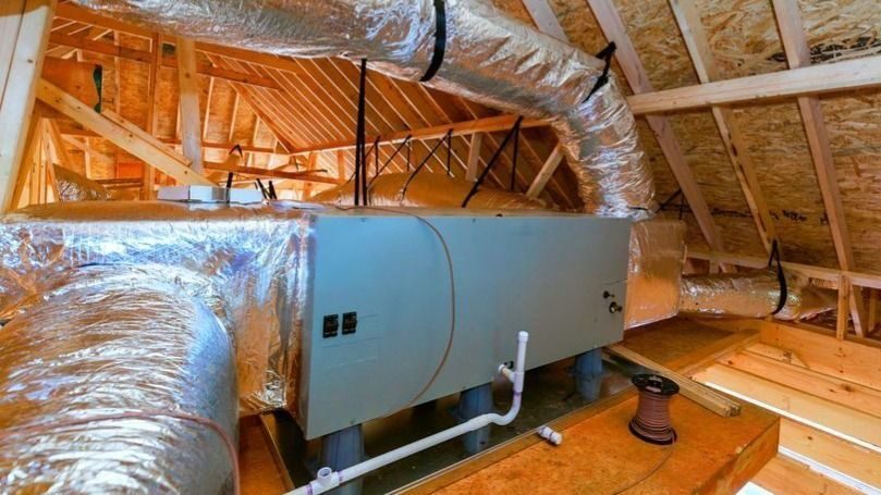 Attic view of an HVAC air handler with insulated ducts and plumbing pipes among wooden rafters.