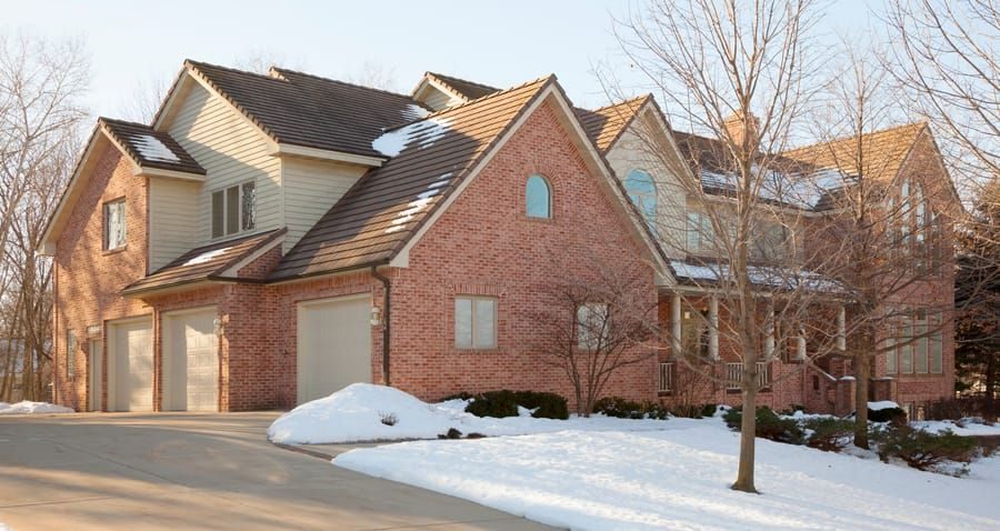 Brick two-story house in winter with a snow-covered yard and driveway, leafless trees nearby