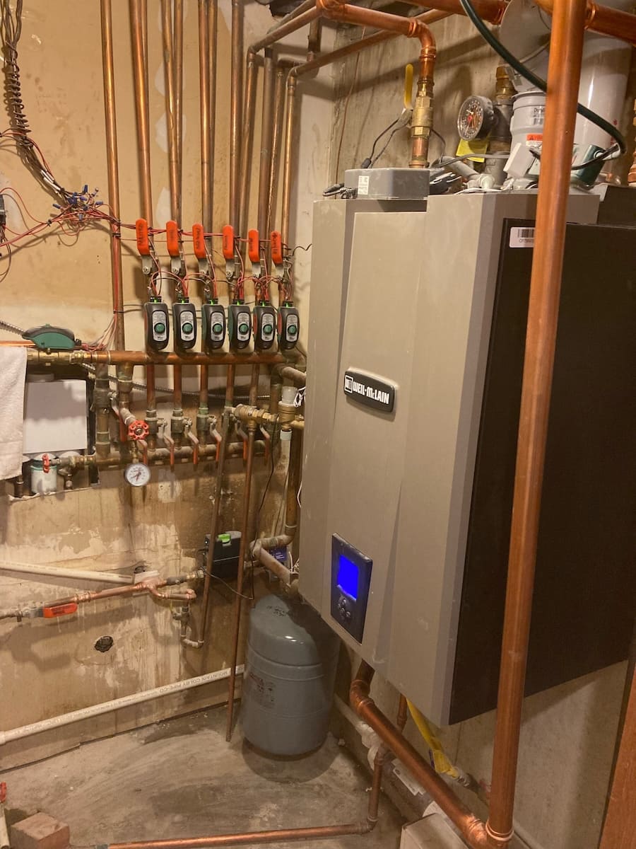 Boiler room with copper pipes, multiple valve handles, and a large wall-mounted boiler with a blue digital display.